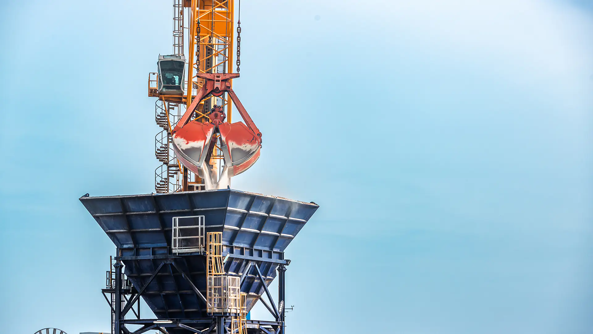 Crane unloading raw sugar into a hopper at a sugar manufacturing facility under a clear blue sky.