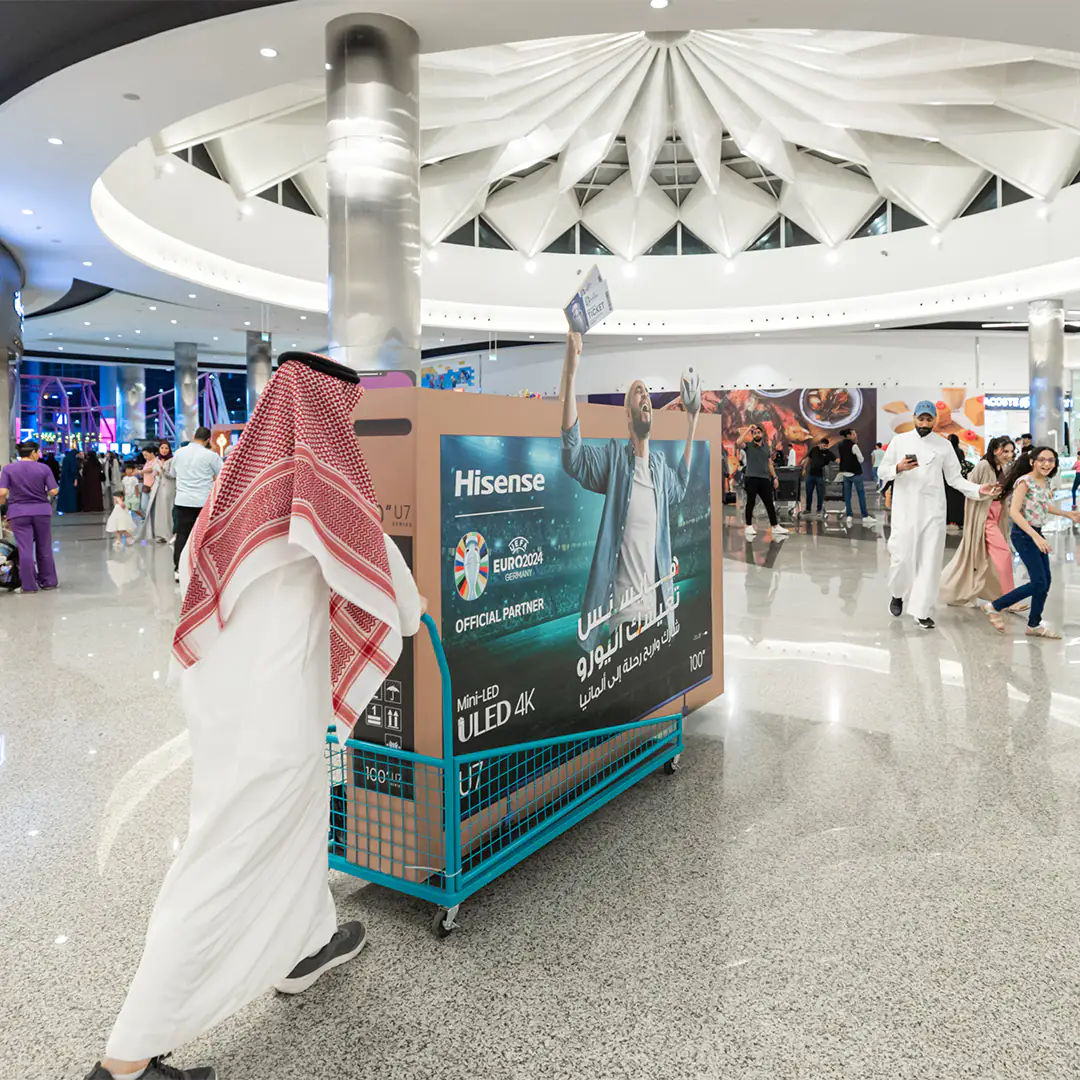 Man in traditional Saudi attire pushing a cart with a Hisense ULED 4K 100-inch TV in a mall, promoting UEFA Euro 2024 partnership