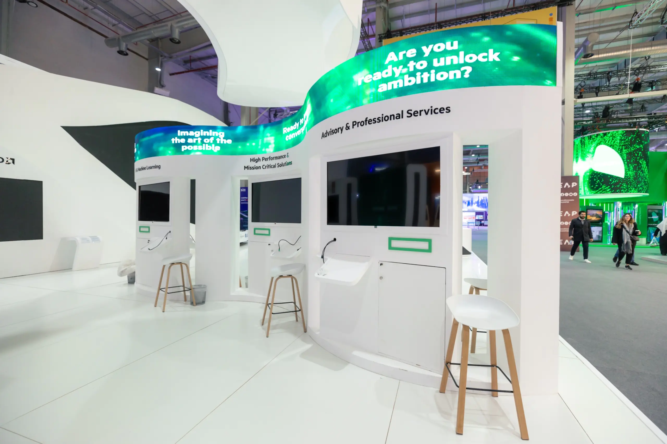 A white trade show booth with interactive displays, bar stools, and curved LED screens with green digital animations.