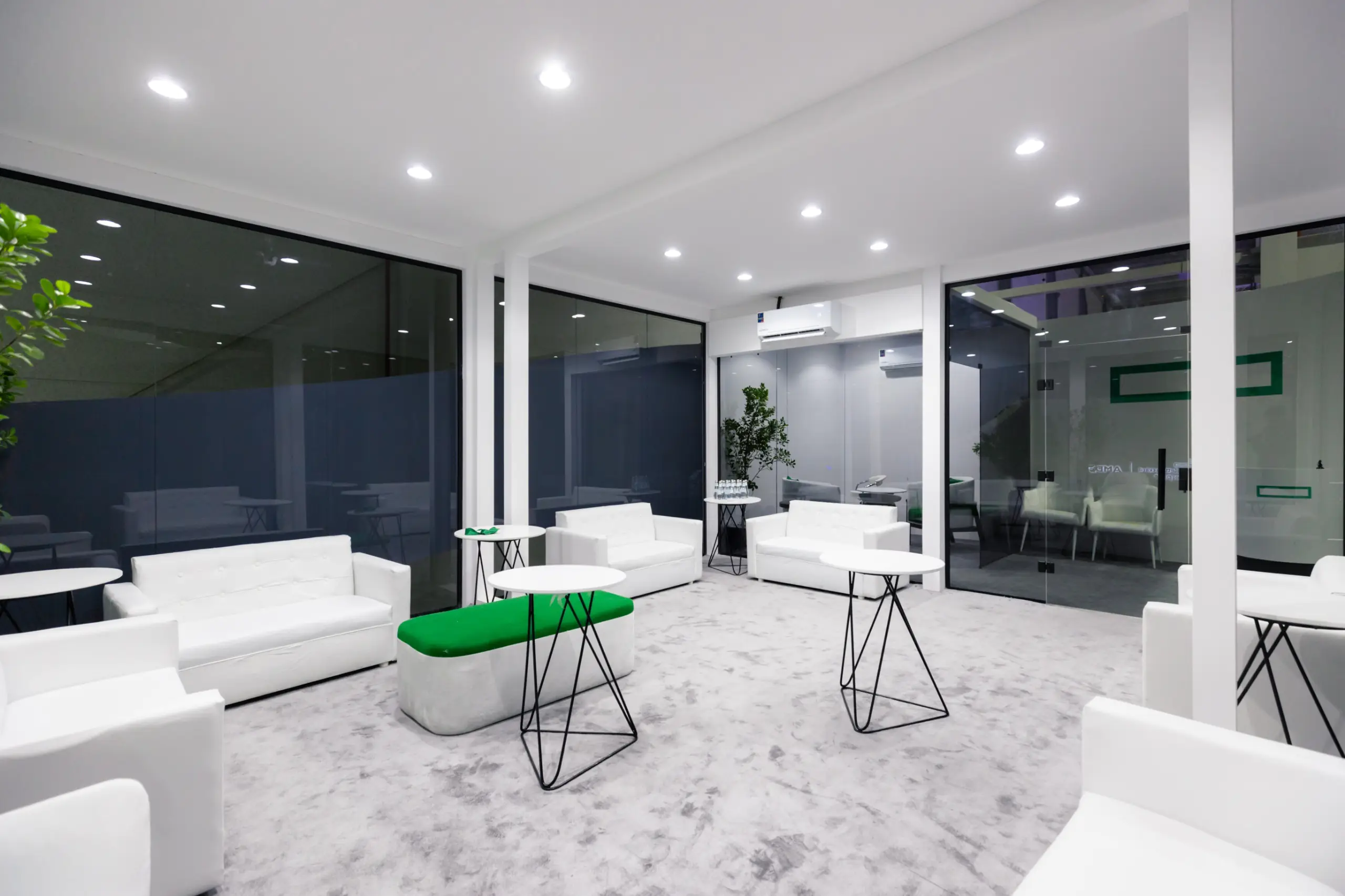 A modern lounge area with white furniture, black wireframe tables, and green accents, enclosed by glass walls.