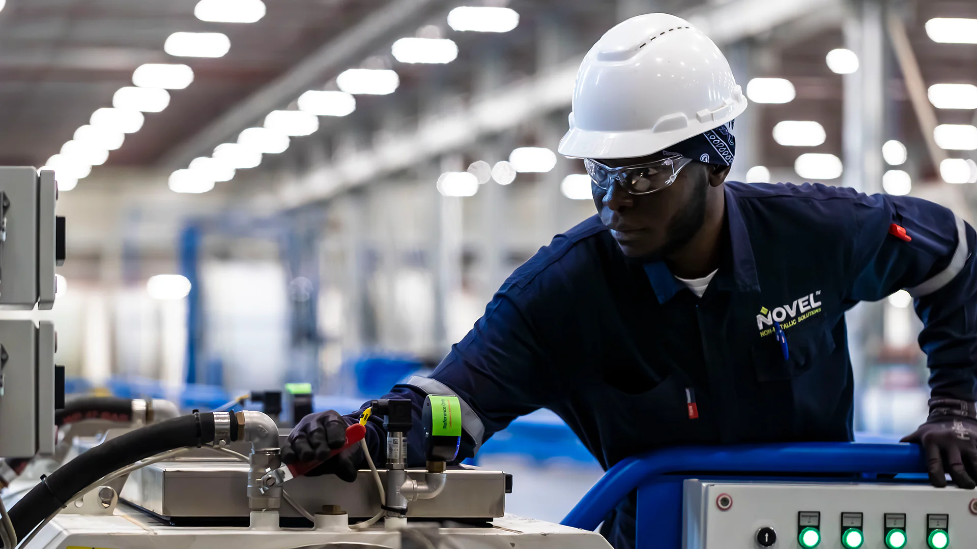 Factory technician in hard hat leans over controls to inspect machinery in a spacious plant