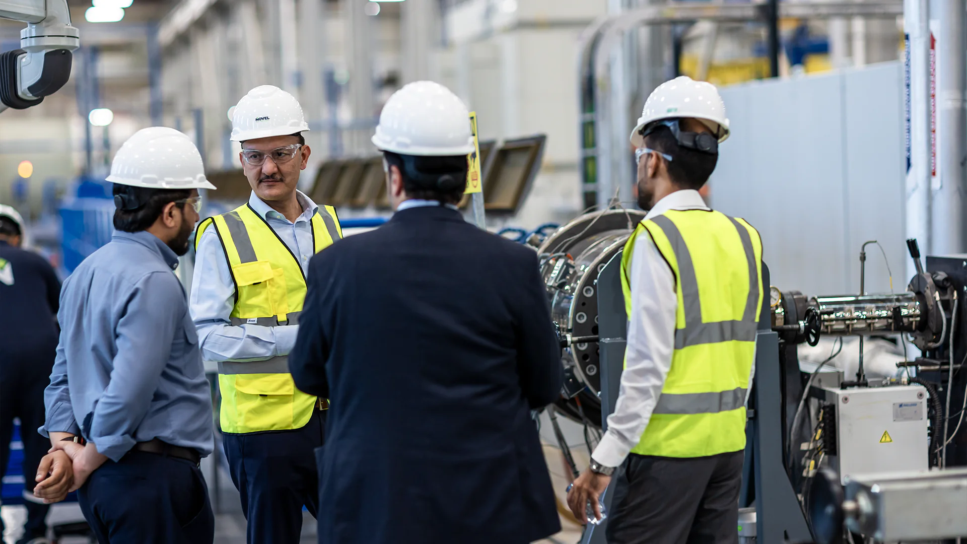 Four factory workers in hard hats and high-vis vests stand discussing near machinery.