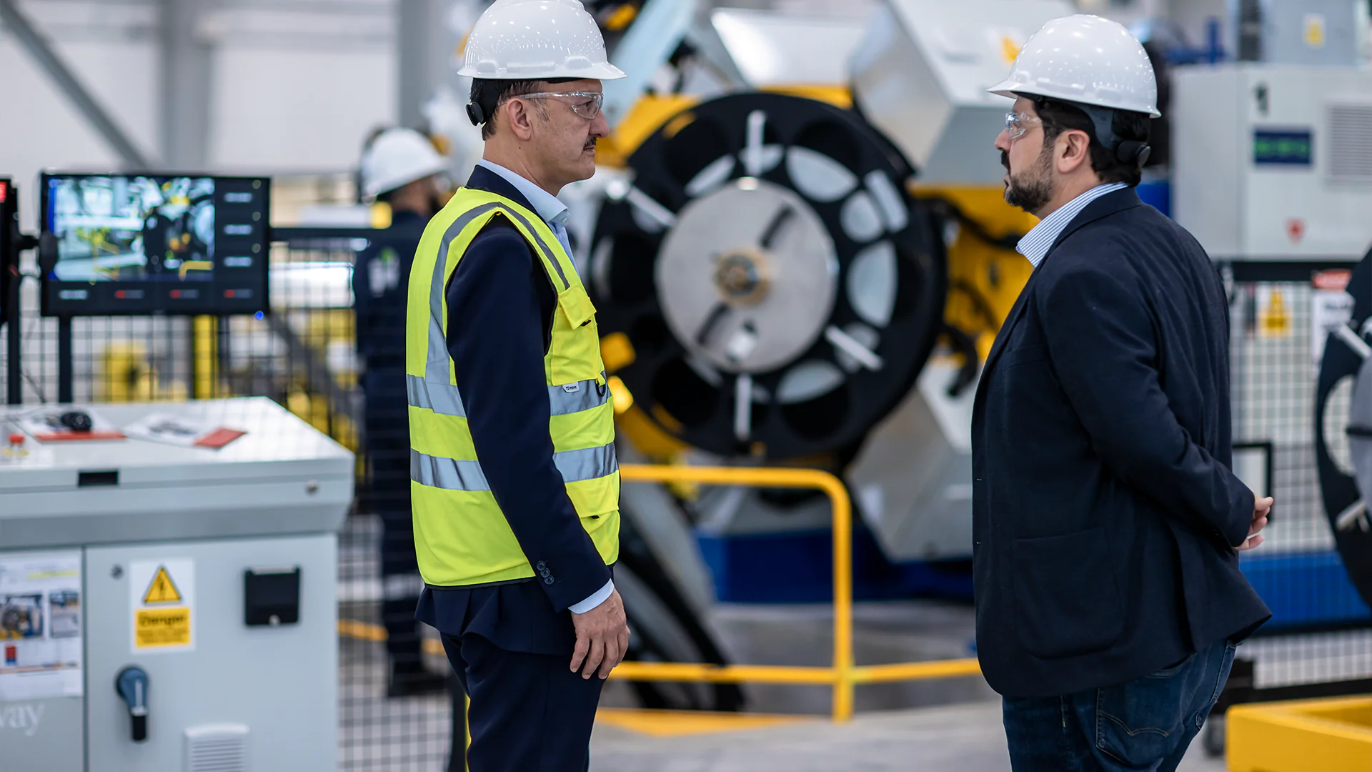 Two men—one in a hi-vis vest—stand facing each other by a machine’s control panel in a factory.