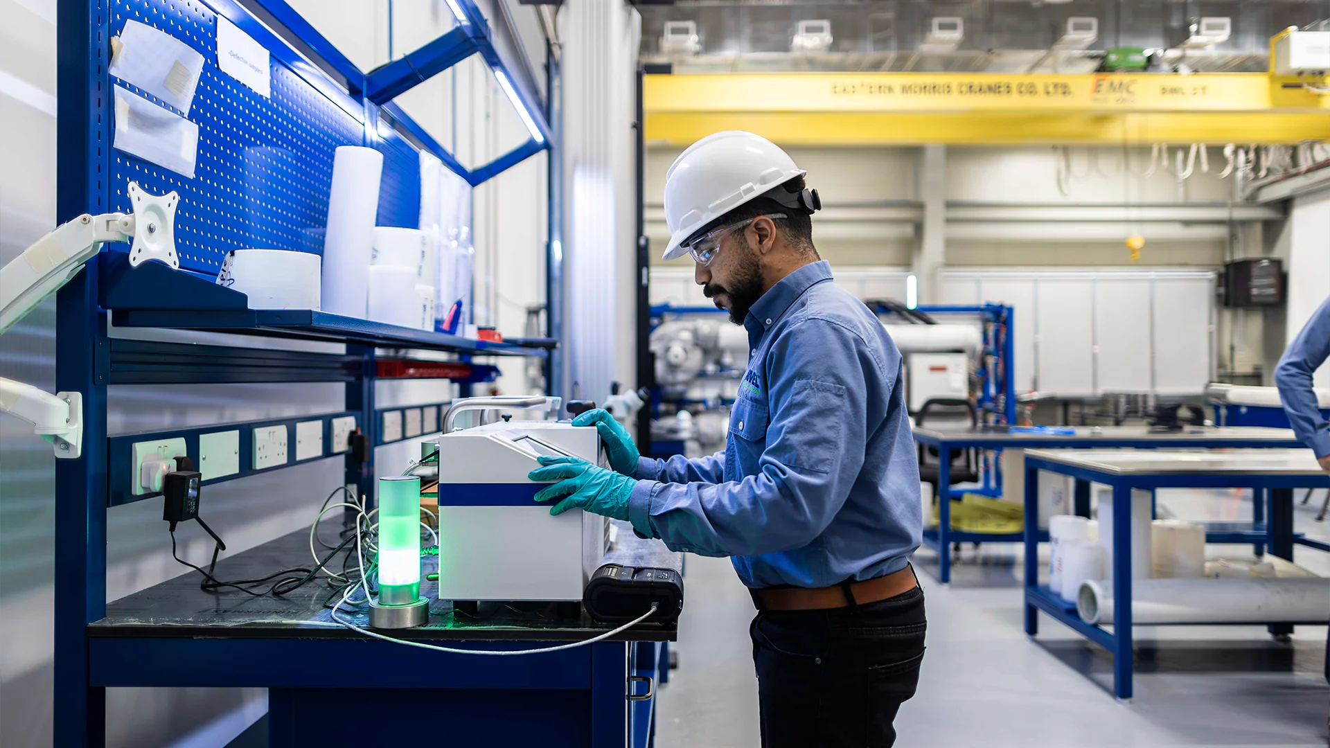 Worker in hard hat and gloves adjusts electronic equipment on a workbench inside a factory.