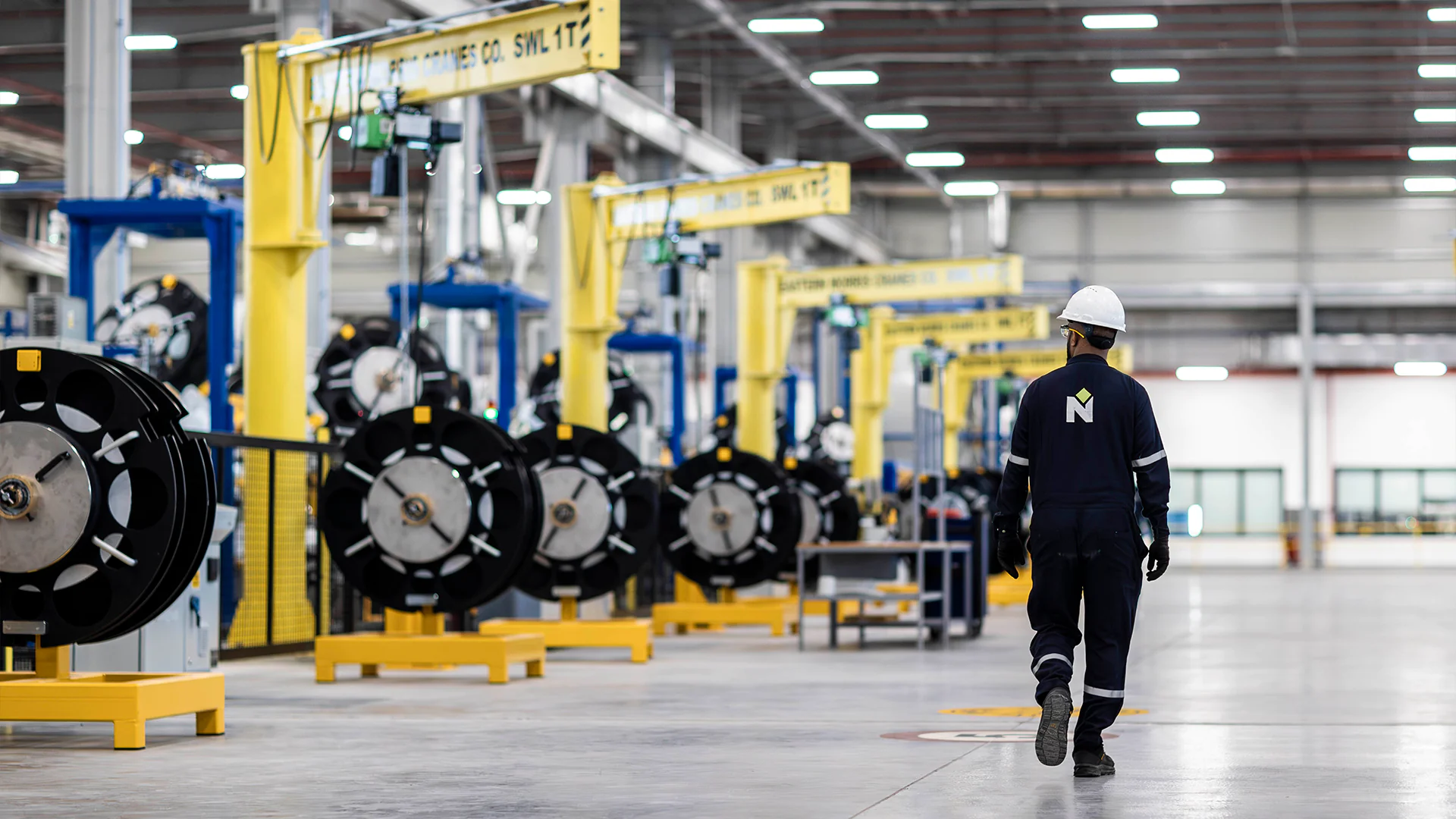 Employee in safety gear walks past rows of cable spools under yellow overhead cranes in a plant.