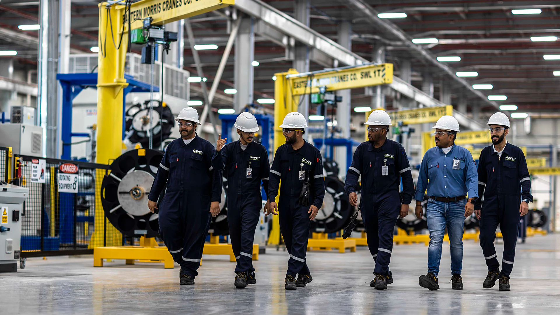 Five workers in hard hats and coveralls walk together through a large industrial workshop.
