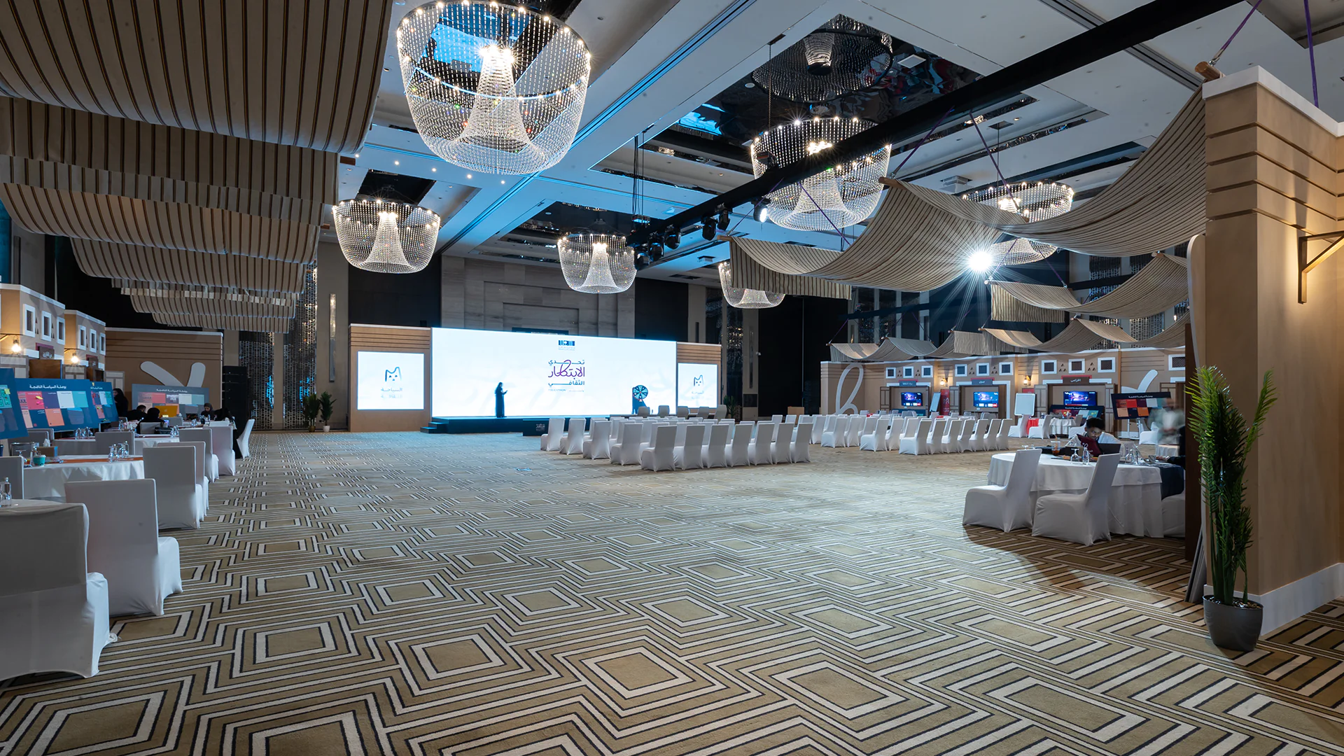 Expansive event hall with chandeliers, display booths, and a stage ready for final Policython presentations.