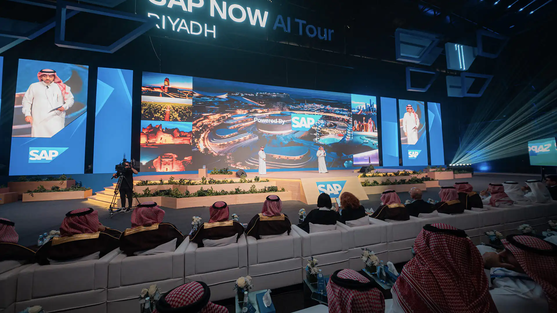 SAP NOW AI Tour keynote stage showing Saudi landmarks and speakers presenting.