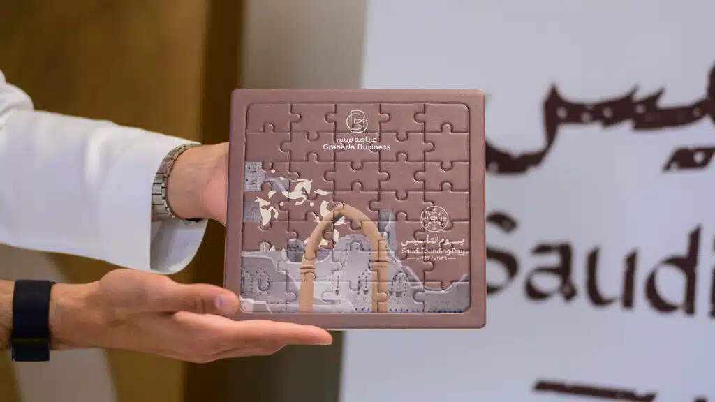 A person holding a Granada Business branded puzzle featuring Saudi heritage imagery for Founding Day